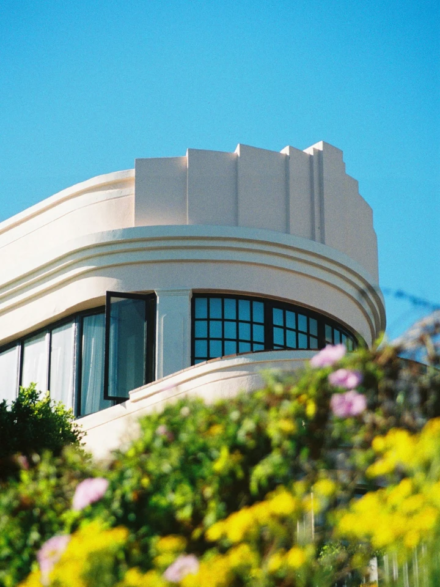Architecture Art Déco blanche aux lignes courbes, fenêtres noires, ciel bleu vif et fleurs colorées en premier plan flou.