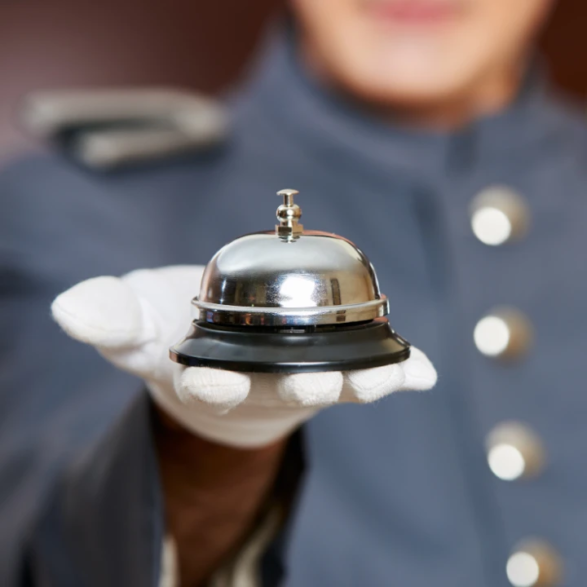 Gant blanc tenant une cloche d’hôtel, uniforme bleu en arrière-plan flou — symbole de service et d’accueil.