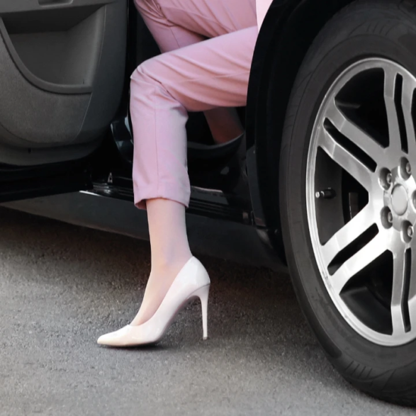 Jambes en pantalon rose et escarpins blancs sortant d’une voiture noire, sur asphalte. Détail élégant et urbain.