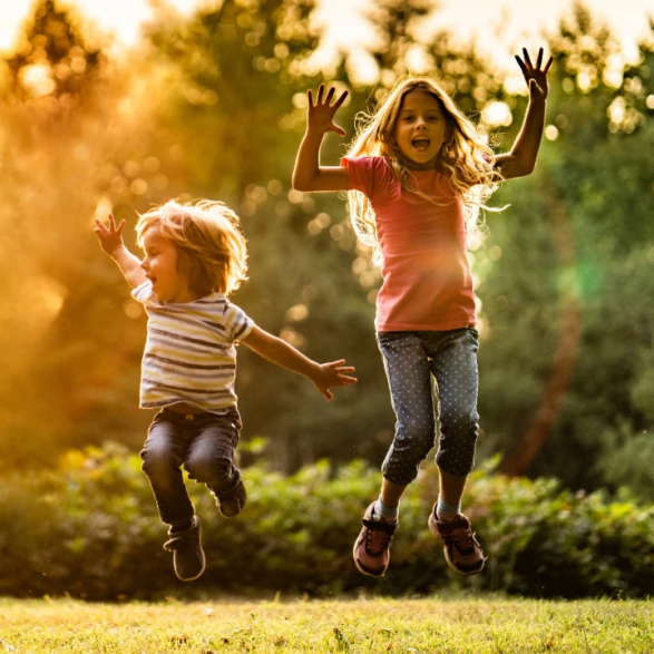 Deux enfants, garçon et fille, sautent en l’air dans un parc ensoleillé. Leurs bras levés expriment la joie pure, baignés d’une lumière dorée chaude.