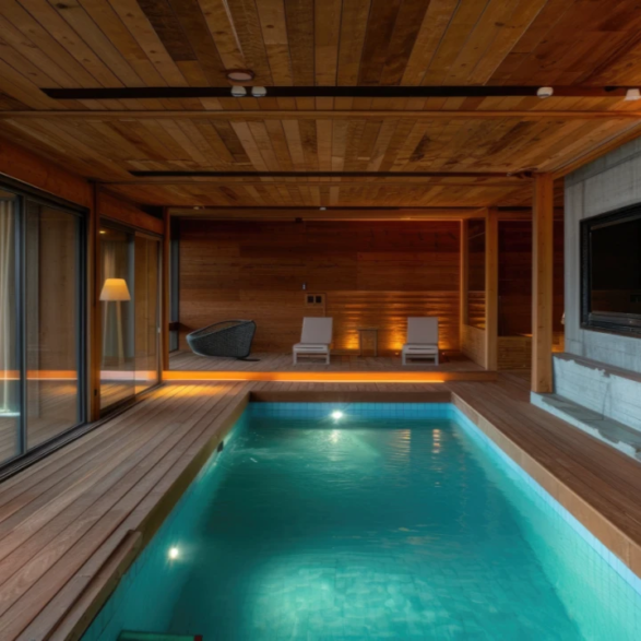 Piscine intérieure luxueuse avec terrasse en bois, chaises longues et éclairage tamisé chaleureux.